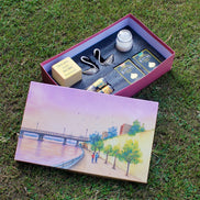An open pink gift box displayed on grass, containing a small white candle, a silver-toned metal spoon, two packaged items with gold and blue labels, and a yellow soap bar. The lid rests beside it and features a painted scene of a riverside walkway with trees and a bridge.