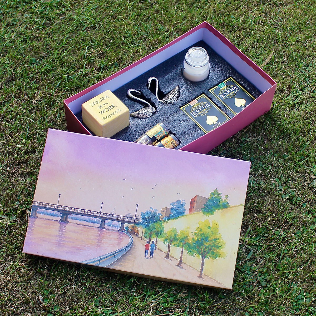 An open pink gift box displayed on grass, containing a small white candle, a silver-toned metal spoon, two packaged items with gold and blue labels, and a yellow soap bar. The lid rests beside it and features a painted scene of a riverside walkway with trees and a bridge.