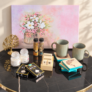 A styled tabletop arrangement featuring a large pastel floral painting in the background, with mugs, candles, spice jars, a wooden calendar, decorative boxes, and a gold tree ornament placed on a black marble table.