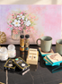 A styled tabletop arrangement featuring a large pastel floral painting in the background, with mugs, candles, spice jars, a wooden calendar, decorative boxes, and a gold tree ornament placed on a black marble table.
