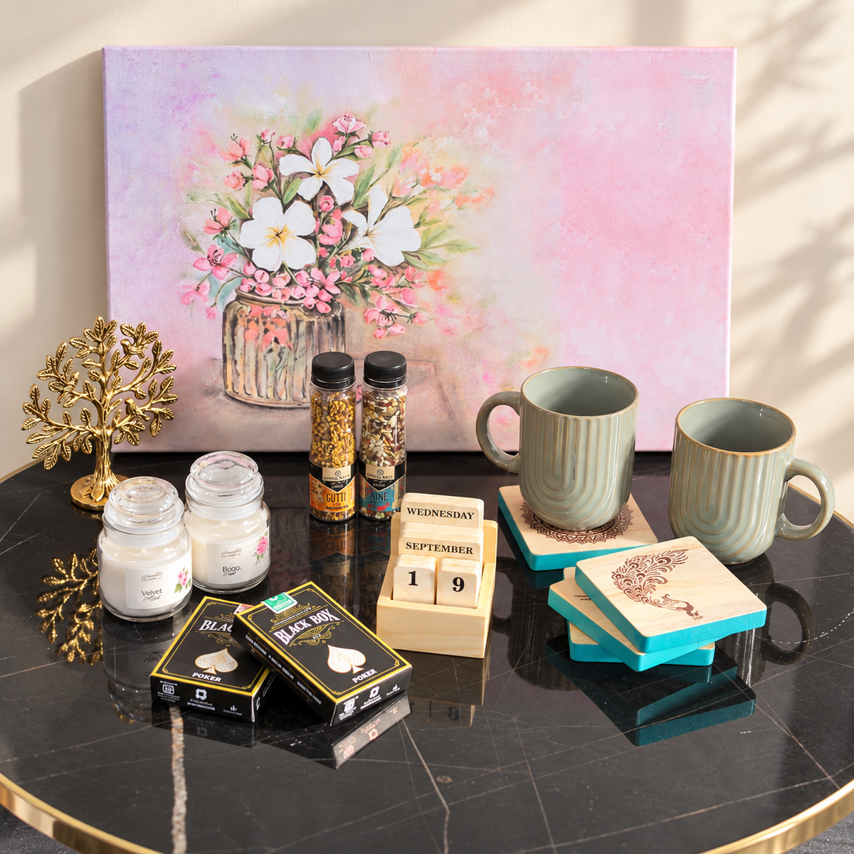A styled tabletop arrangement featuring a large pastel floral painting in the background, with mugs, candles, spice jars, a wooden calendar, decorative boxes, and a gold tree ornament placed on a black marble table.