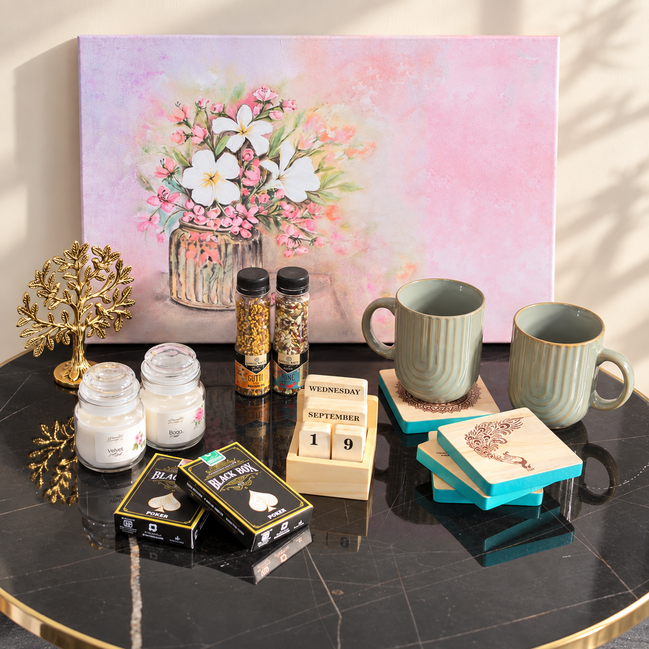 A styled tabletop arrangement featuring a large pastel floral painting in the background, with mugs, candles, spice jars, a wooden calendar, decorative boxes, and a gold tree ornament placed on a black marble table.