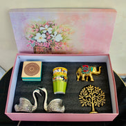 An open pink gift box with a floral illustration on the inside lid, containing four decorative items: two silver swan figurines, a green and yellow patterned cup, a small gold elephant, and a gold tree of life ornament, all arranged on a dark insert.