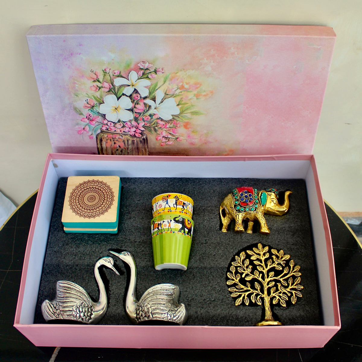 An open pink gift box with a floral illustration on the inside lid, containing four decorative items: two silver swan figurines, a green and yellow patterned cup, a small gold elephant, and a gold tree of life ornament, all arranged on a dark insert.