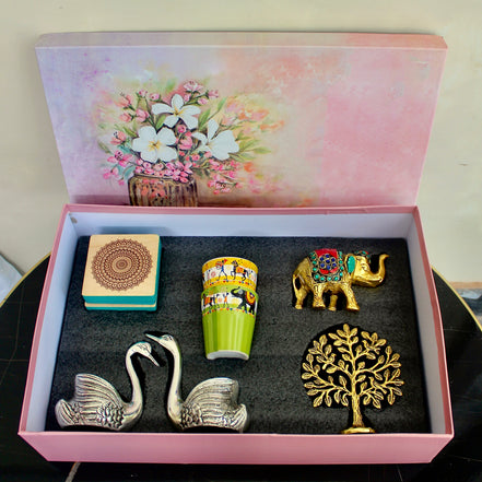 An open pink gift box with a floral illustration on the inside lid, containing four decorative items: two silver swan figurines, a green and yellow patterned cup, a small gold elephant, and a gold tree of life ornament, all arranged on a dark insert.