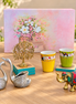 A styled tabletop display featuring a pink floral gift box, two yellow and green patterned cups on teal coasters, a gold elephant figurine, a pair of silver swan-shaped objects, and a gold tree sculpture, all arranged on a wooden surface with a blurred outdoor garden background.