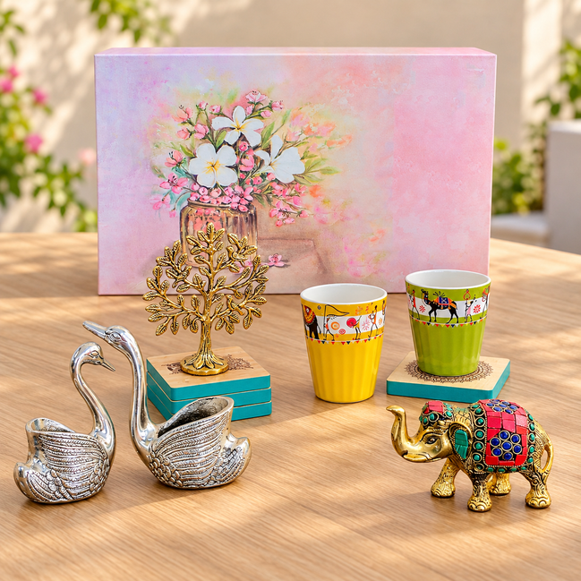 A styled tabletop display featuring a pink floral gift box, two yellow and green patterned cups on teal coasters, a gold elephant figurine, a pair of silver swan-shaped objects, and a gold tree sculpture, all arranged on a wooden surface with a blurred outdoor garden background.