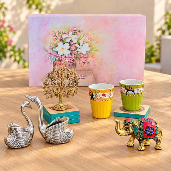 A styled tabletop display featuring a pink floral gift box, two yellow and green patterned cups on teal coasters, a gold elephant figurine, a pair of silver swan-shaped objects, and a gold tree sculpture, all arranged on a wooden surface with a blurred outdoor garden background.