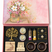 A pink gift box set with a floral lid illustration and an open black tray containing a gold tree ornament, two small candles, two spice jars, two black-and-gold spade boxes, a wooden peacock stamp, a wooden calendar block, and two dark brown cups.