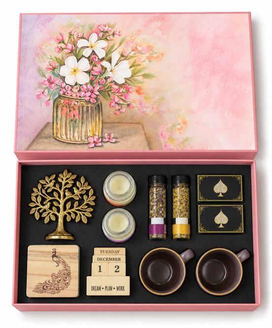 A pink gift box set with a floral lid illustration and an open black tray containing a gold tree ornament, two small candles, two spice jars, two black-and-gold spade boxes, a wooden peacock stamp, a wooden calendar block, and two dark brown cups.