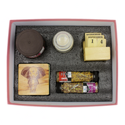 A boxed gift set arranged in a pink-lined tray with gray foam insert. The set includes a small jar with a dark lid, a small clear jar, a wooden calendar block showing Wednesday 14 September, a square wooden coaster with an elephant engraving, two spice jars with labels, and a small round item in the center.