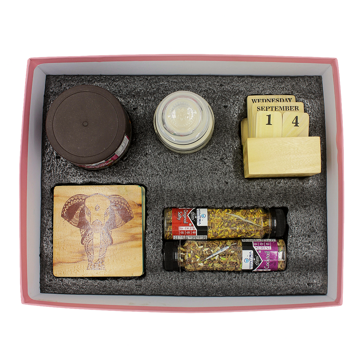 A boxed gift set arranged in a pink-lined tray with gray foam insert. The set includes a small jar with a dark lid, a small clear jar, a wooden calendar block showing Wednesday 14 September, a square wooden coaster with an elephant engraving, two spice jars with labels, and a small round item in the center.